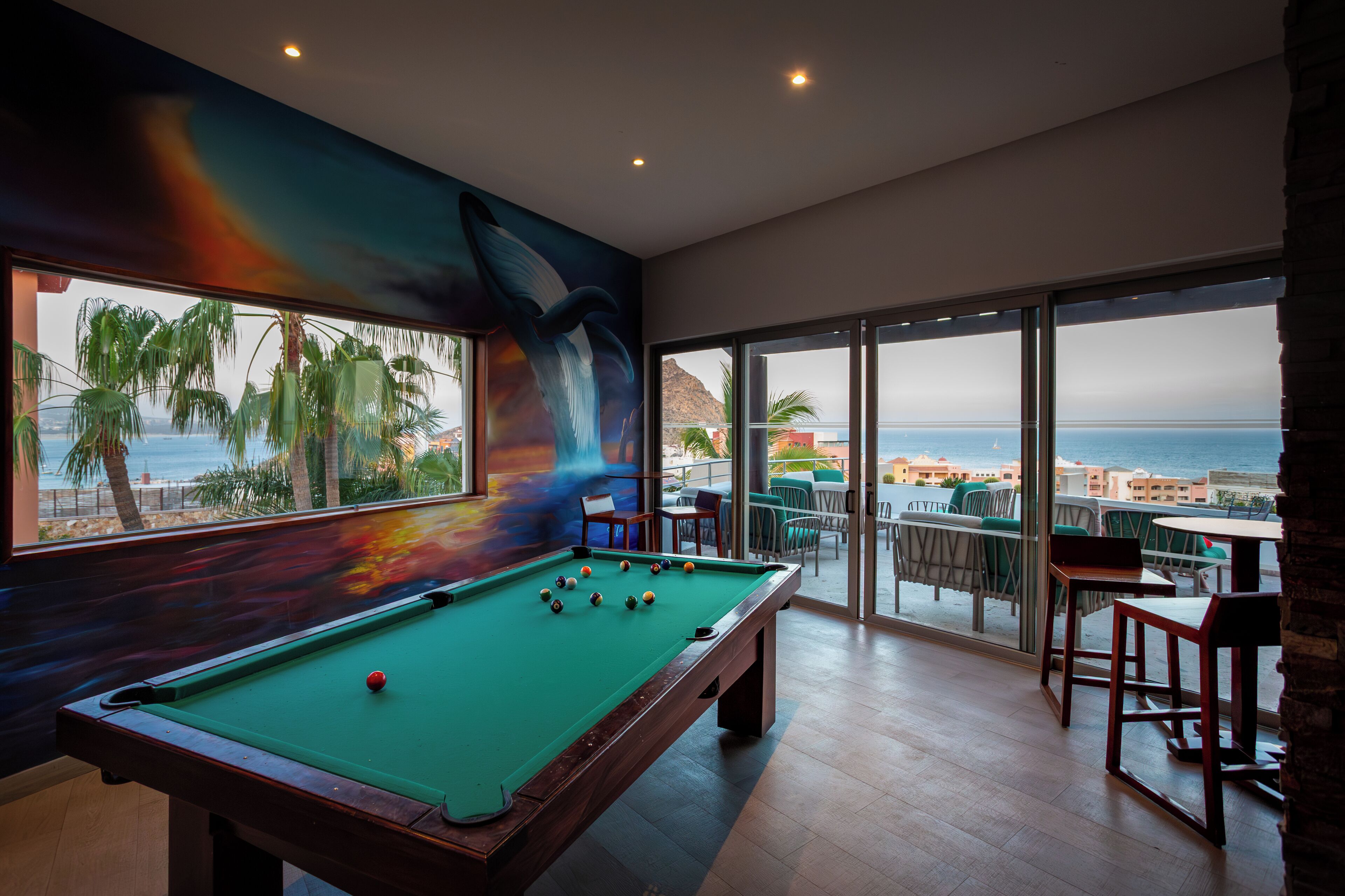 A pool table with a view of the ocean and palm trees through large windows.