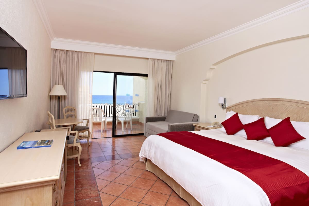 A hotel room with a bed, a desk, a chair, a balcony with a view of the sea, and a television on the wall.