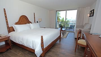 A hotel room with a large wooden four-poster bed, a small desk with a telephone, a chair, a sofa, and a balcony with a view of palm trees.