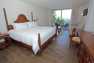 A hotel room with a large wooden four-poster bed, a small desk with a telephone, a chair, a sofa, and a balcony with a view of palm trees.