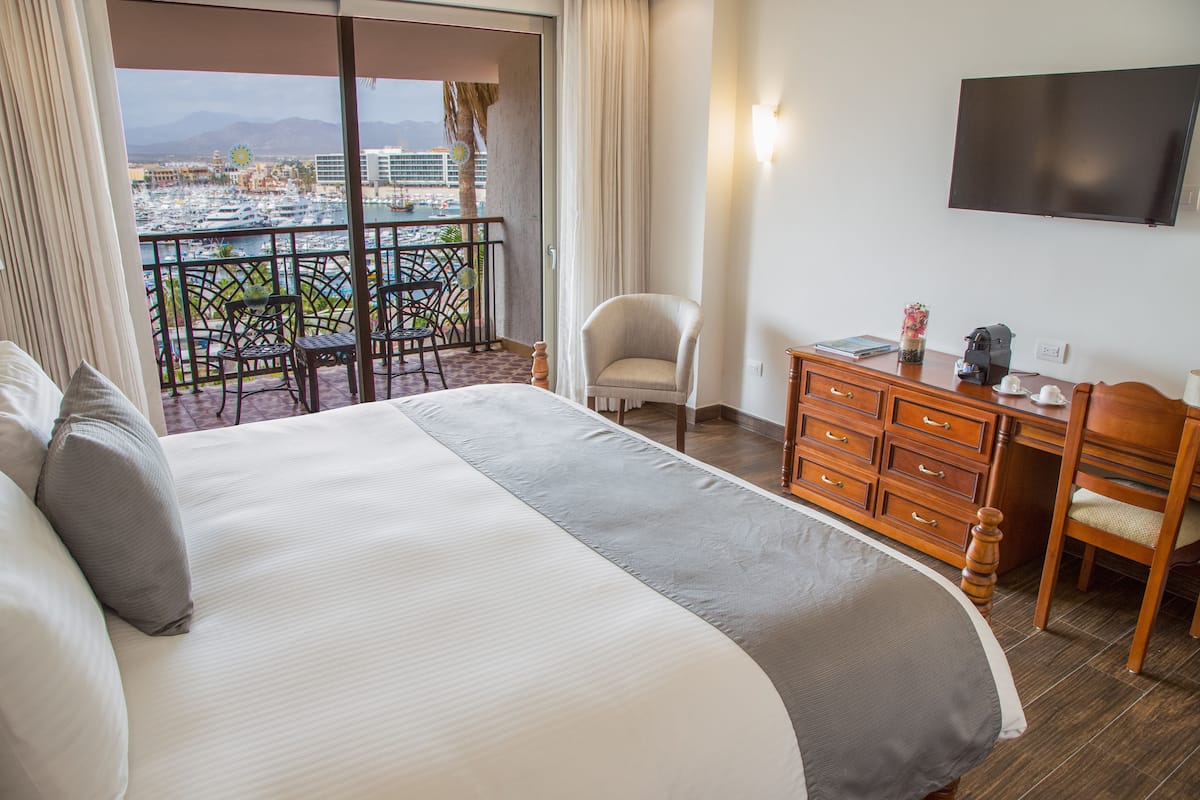 A hotel room with a large bed, a desk with a chair, a TV mounted on the wall, and a balcony with a view of the city and mountains.
