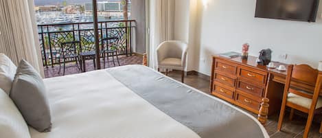 A hotel room with a large bed, a desk with a chair, a TV mounted on the wall, and a balcony with a view of the city and mountains.