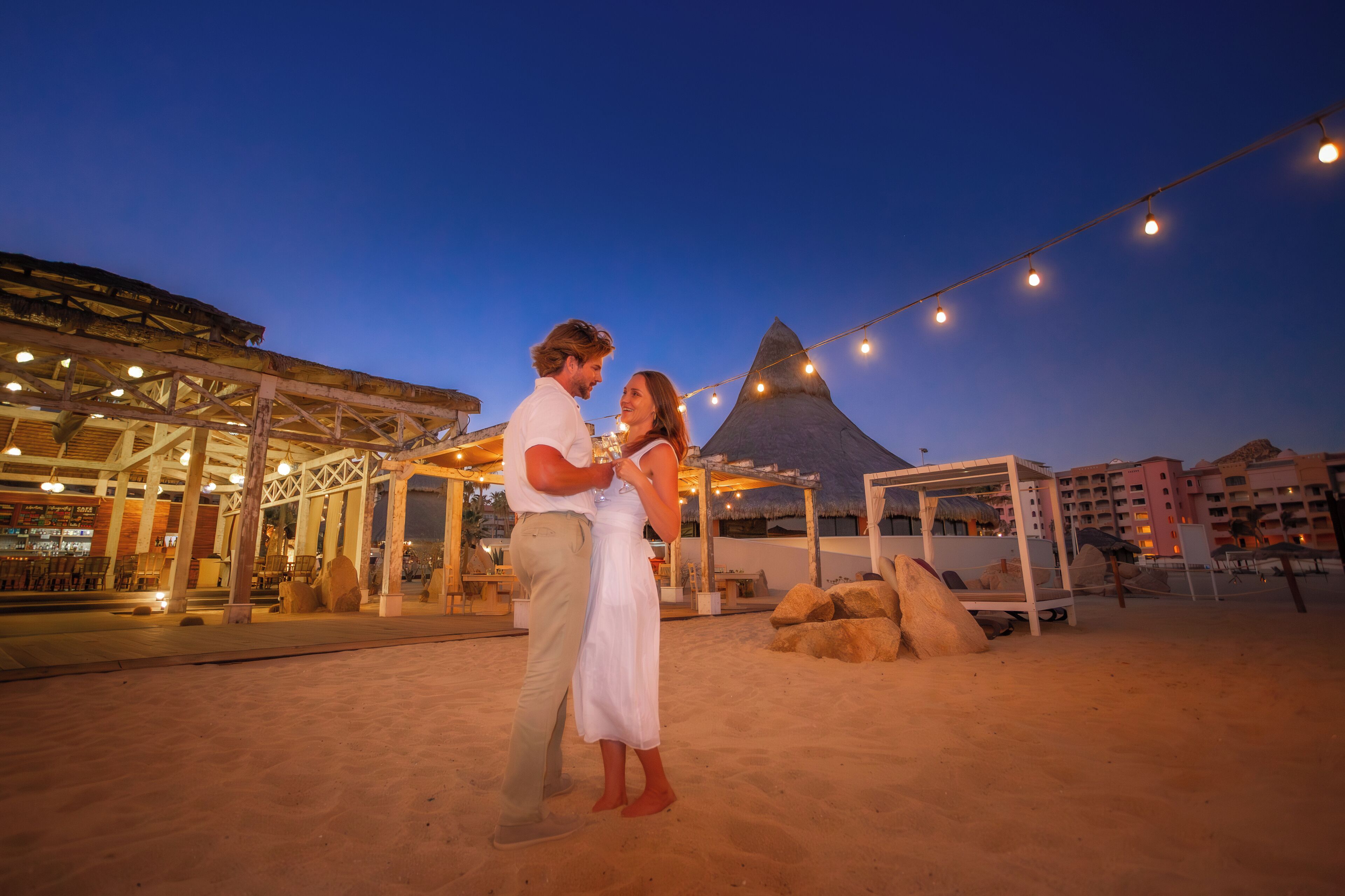 A couple standing on a beach with a thatched-roof structure and string lights in the background.