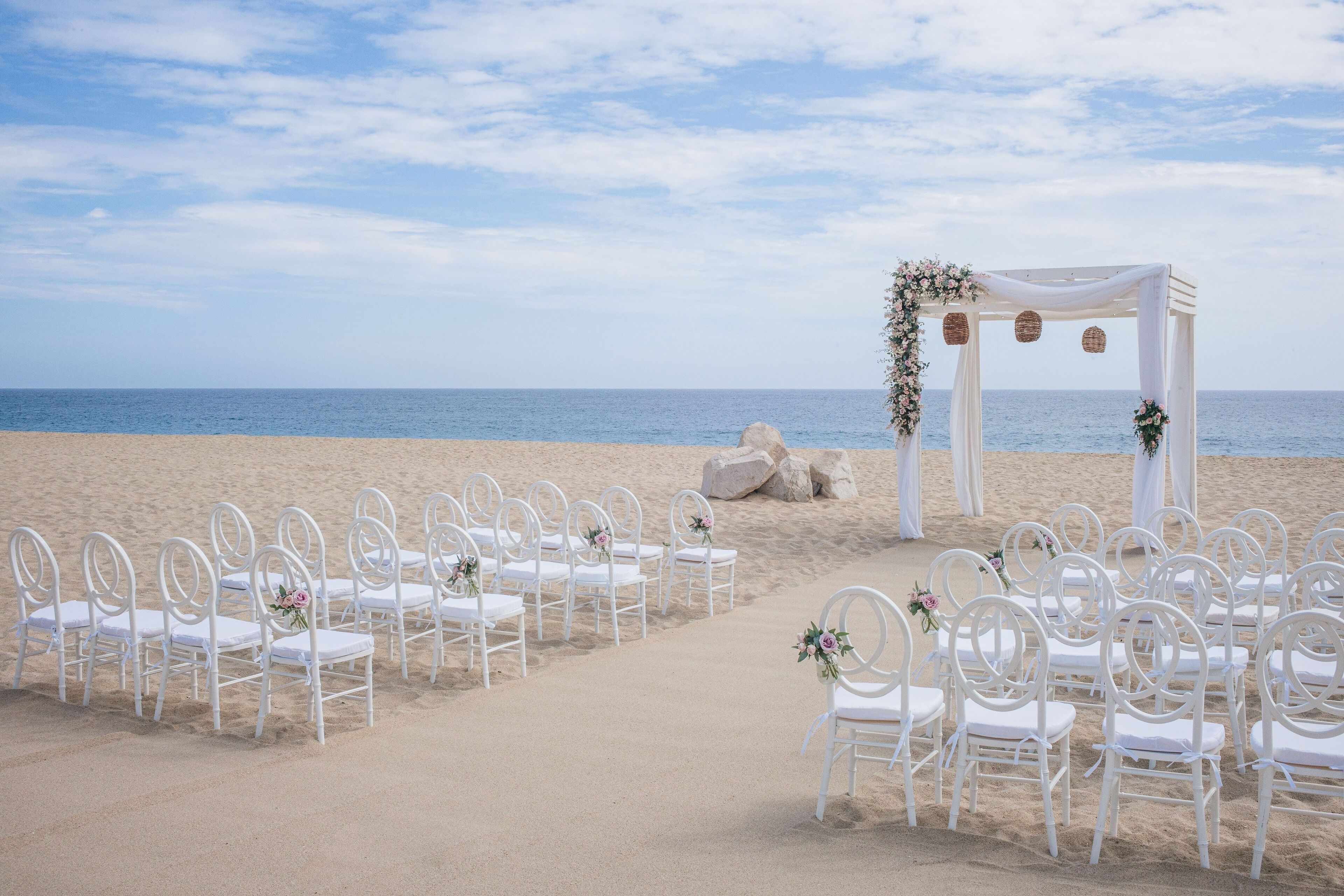 A beach wedding setup with white chairs and a white arch adorned with flowers.