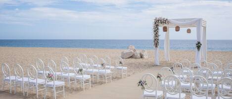 A beach wedding setup with white chairs and a white arch adorned with flowers.