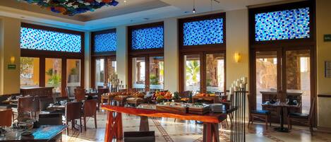A hotel dining area with a buffet, large windows with blue-tinted glass, and a decorative ceiling.