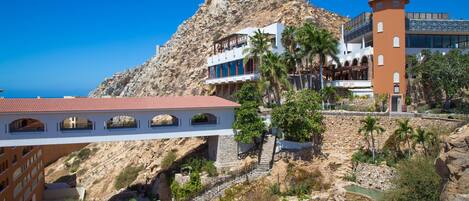 Un hotel frente al mar con un escarpado acantilado al fondo, una escalera de piedra que lleva a los niveles superiores y una zona de piscina.