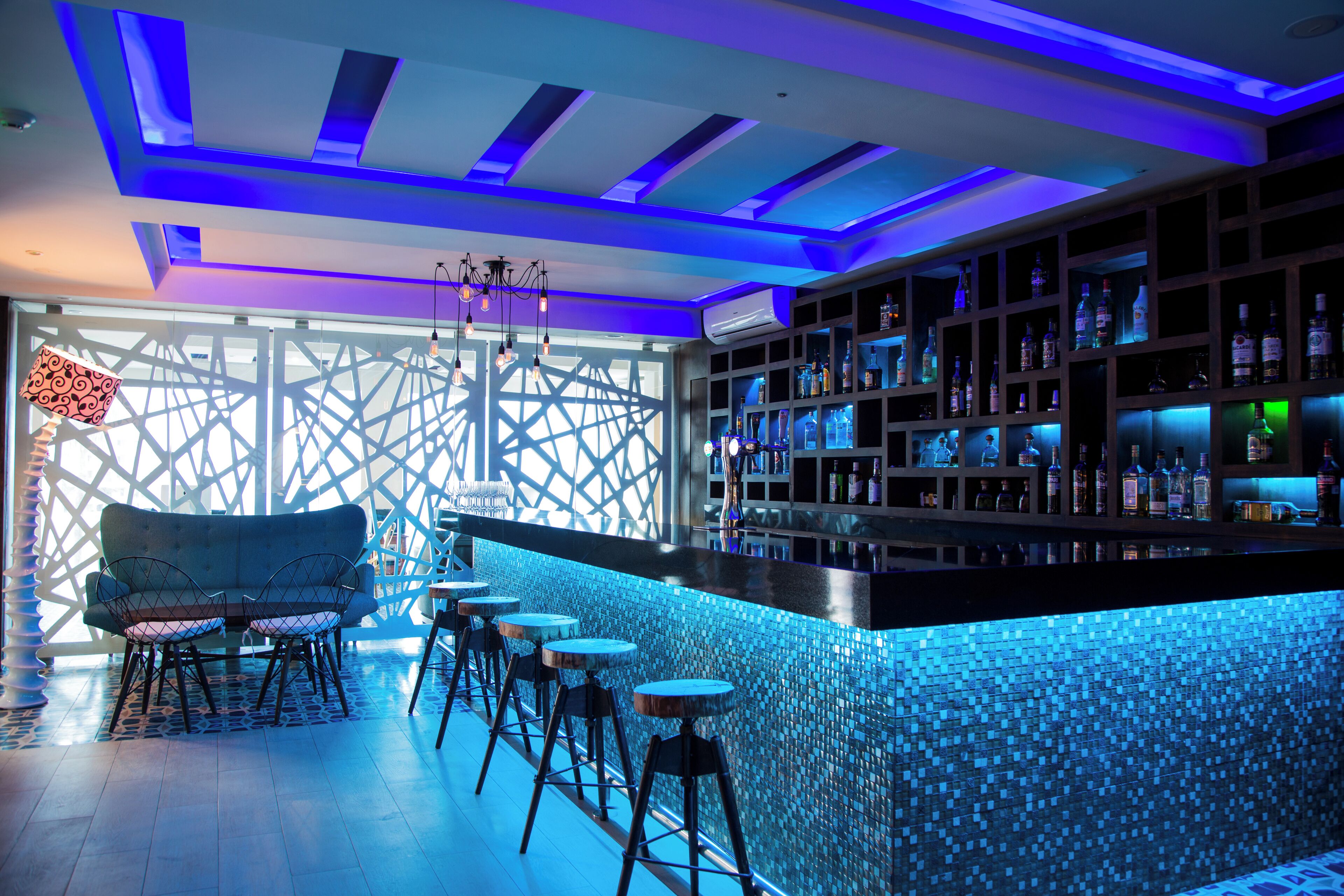 A bar area with a blue-tiled bar counter, a seating area with a sofa and stools, and a patterned wall.