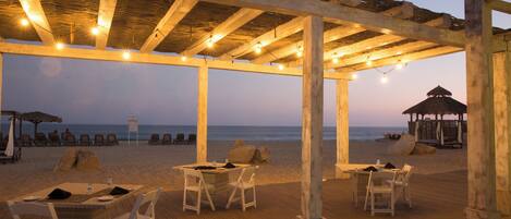 A beachside restaurant with outdoor seating, a wooden deck, and a view of the ocean.