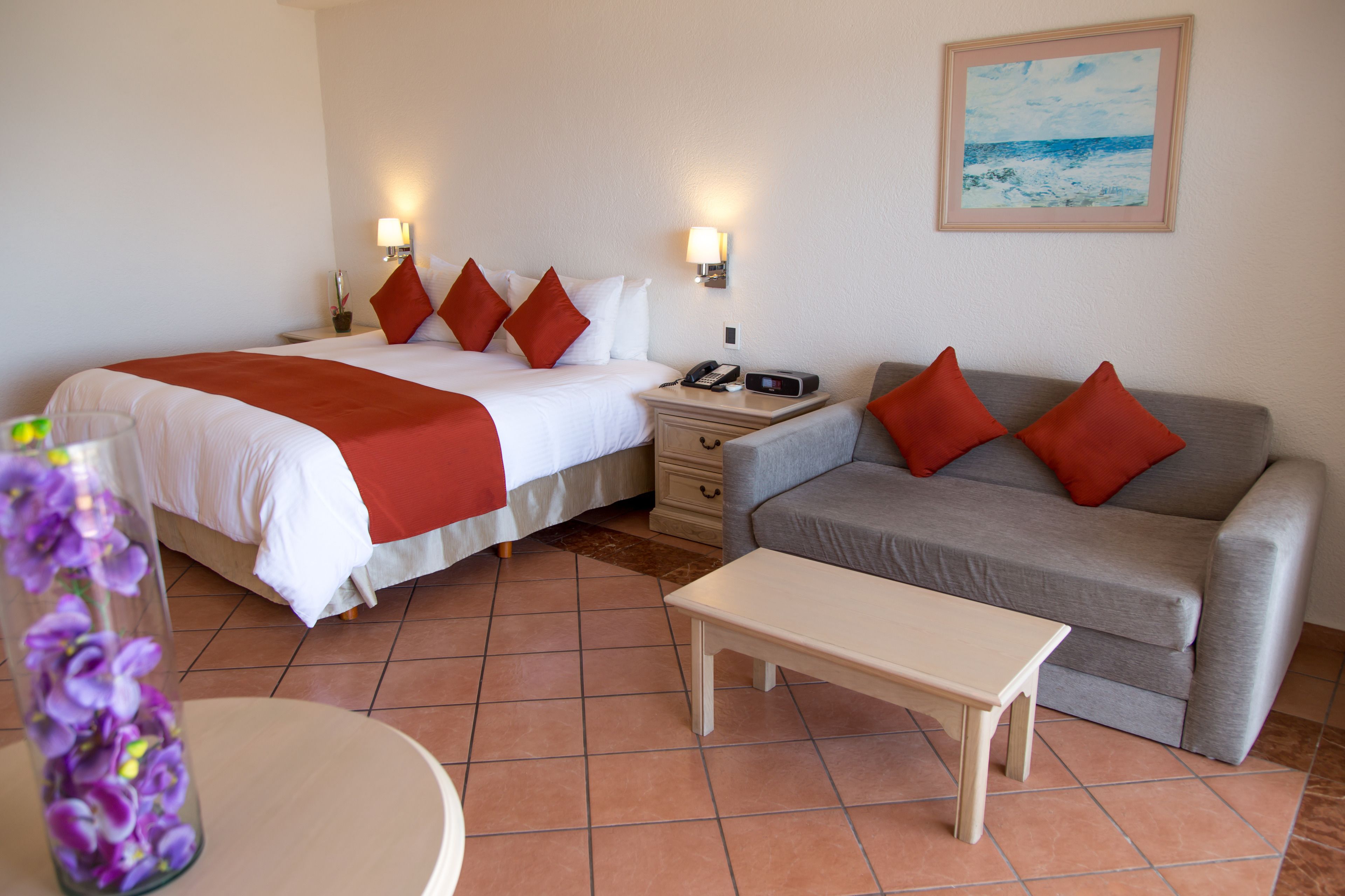 A hotel room with two beds, a grey sofa, a bedside table, and a painting on the wall.