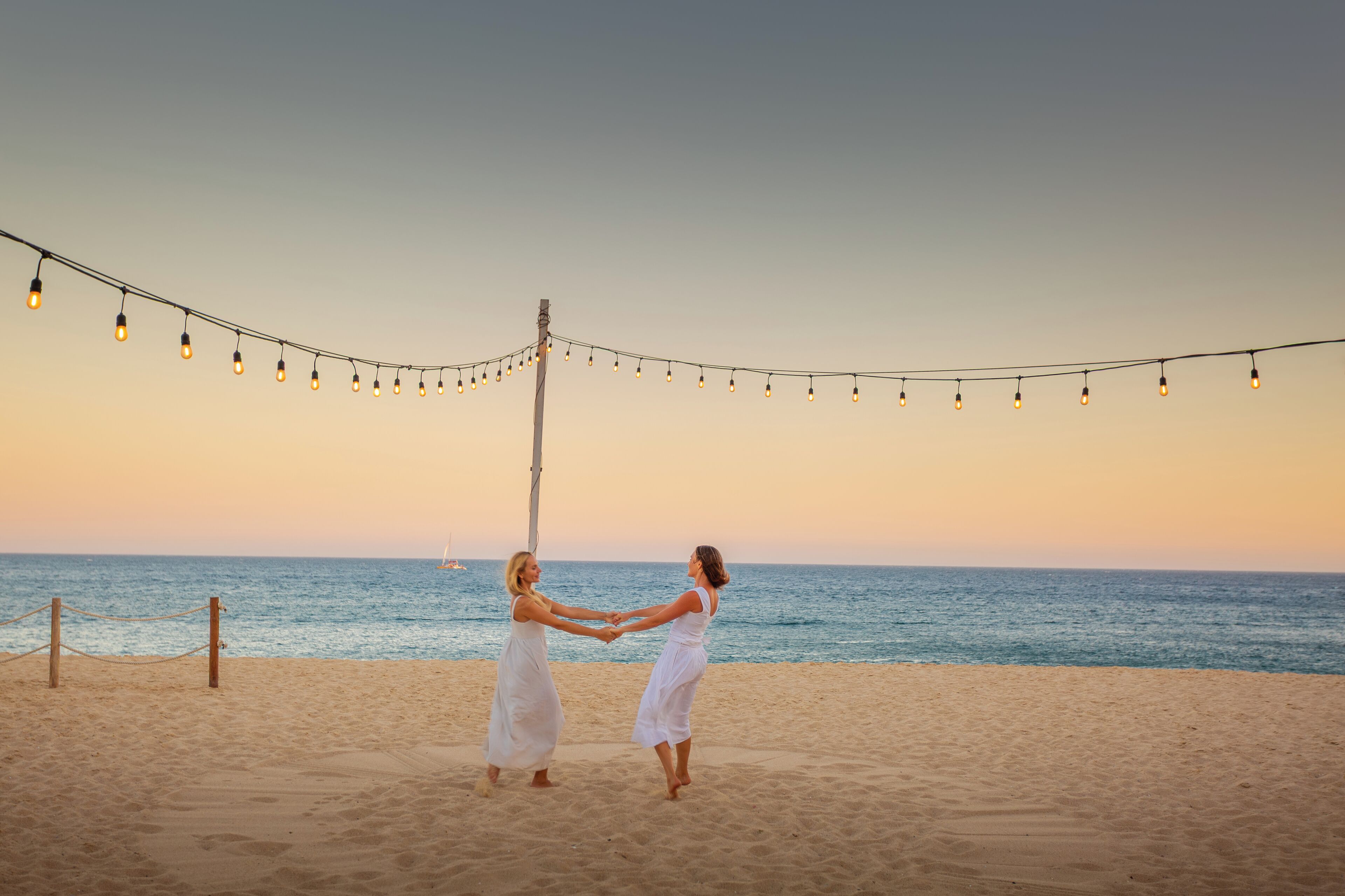 A beach scene with two people holding hands, a string of lights, and a wooden post.