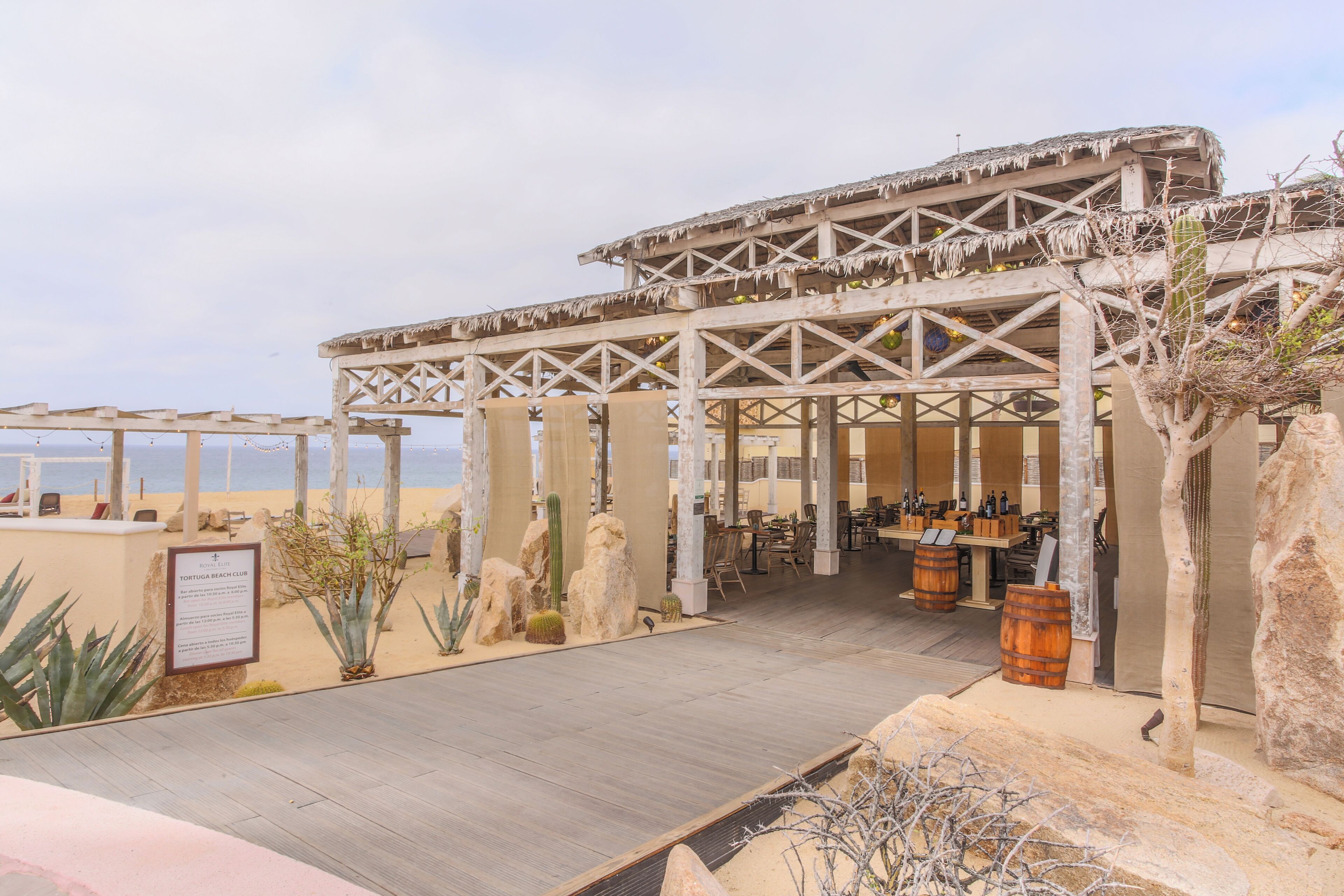 En restaurant ved stranden med udendørs siddepladser, en træpergola og ørkenplanter.