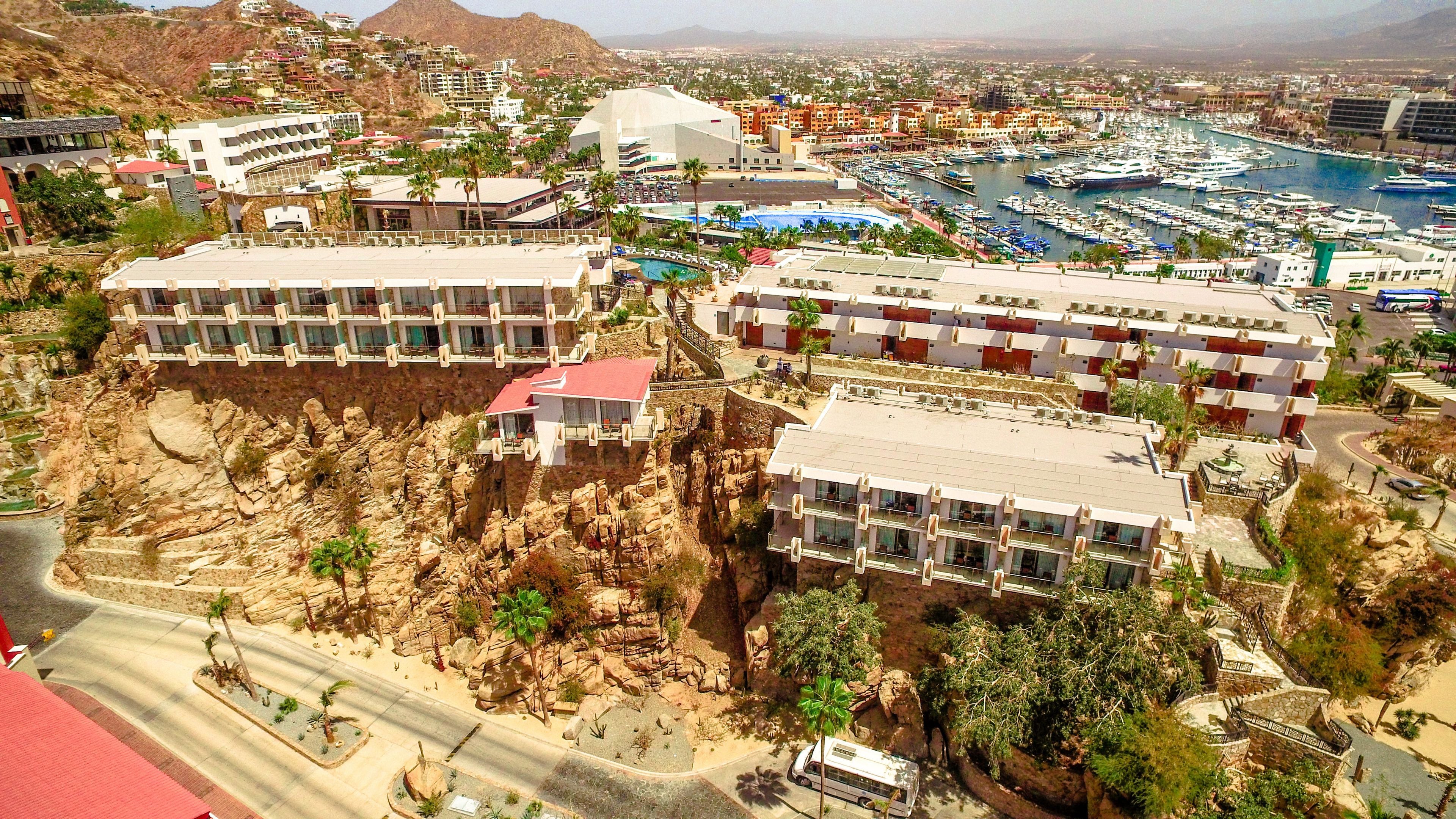 Un hotel frente al mar con varias construcciones, un puerto deportivo y terreno rocoso.