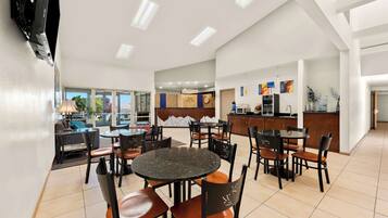 A modern hotel dining area with round tables and chairs, a kitchen in the background, and a view of the outdoors.