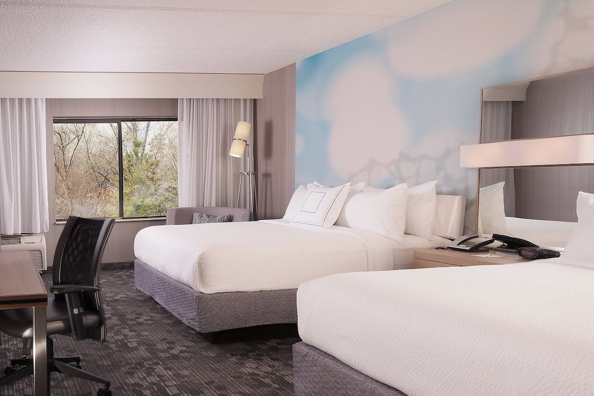 Une chambre d’hôtel avec deux lits, un bureau, une chaise, une lampe et une vue sur les arbres par la fenêtre.