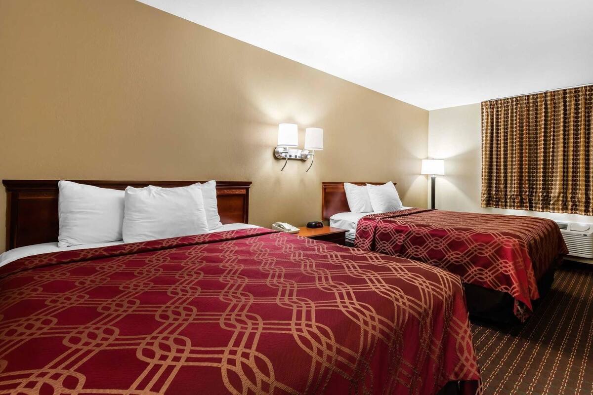 Une chambre d’hôtel avec deux lits, une table de chevet avec un téléphone, une lampe et une fenêtre avec des rideaux.