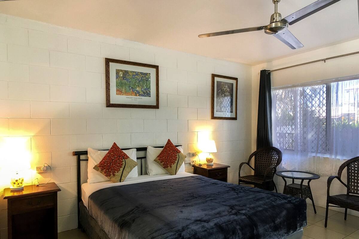 A hotel room with a bed, two nightstands, a ceiling fan, two framed pictures, and a window with a view.