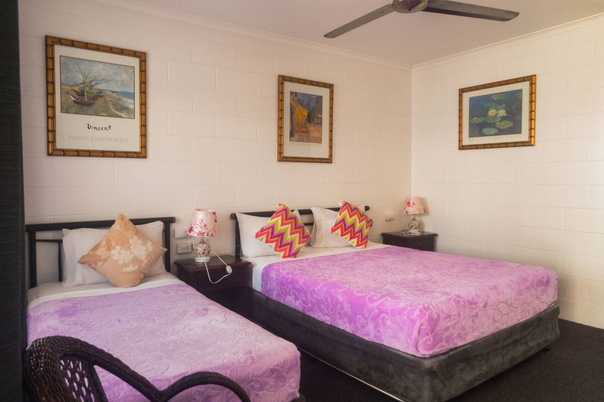 A hotel room with two beds, each with a purple bedspread, a black headboard, and a nightstand with a lamp. There are framed pictures on the wall above the beds.