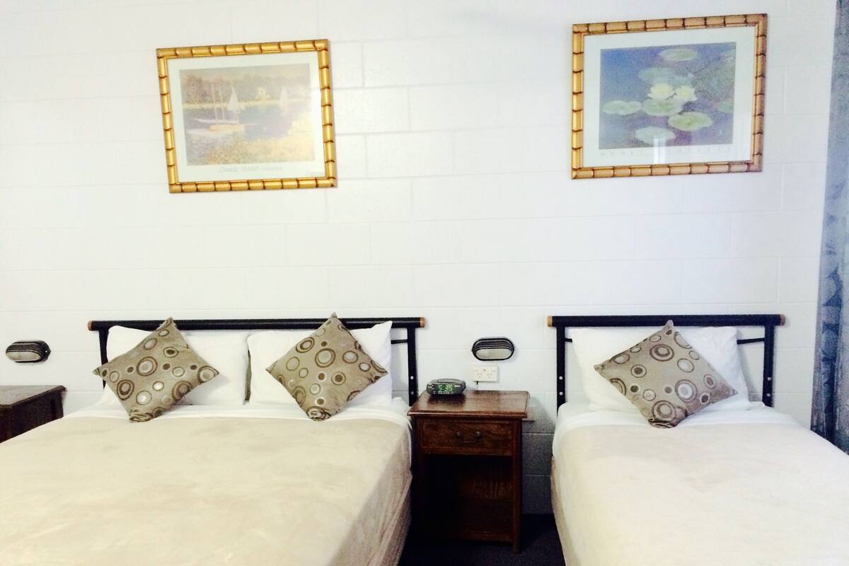 A room with two beds, each with patterned pillows, a bedside table with a clock, and framed pictures on the wall.