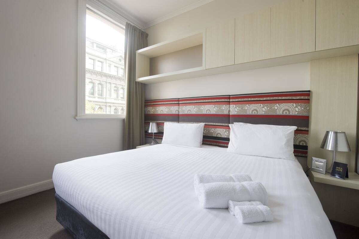 Camera d'albergo con un letto grande, una testiera decorata, una finestra con tende e un comodino con una lampada e un cartello.