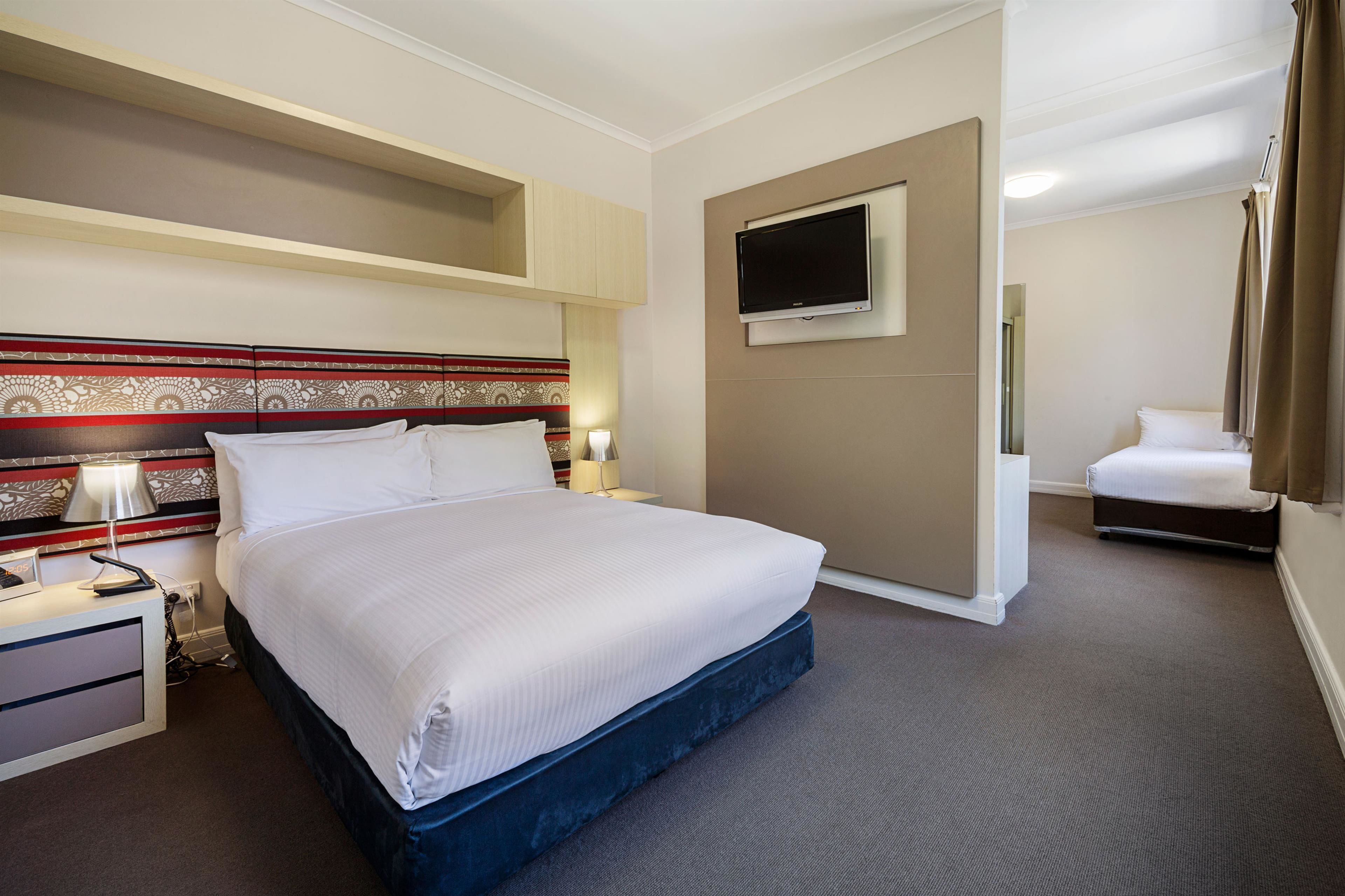 Quarto de hotel com uma cama grande, televisão de tela plana na parede, mesa de cabeceira com abajur e uma pequena escrivaninha com cadeira.
