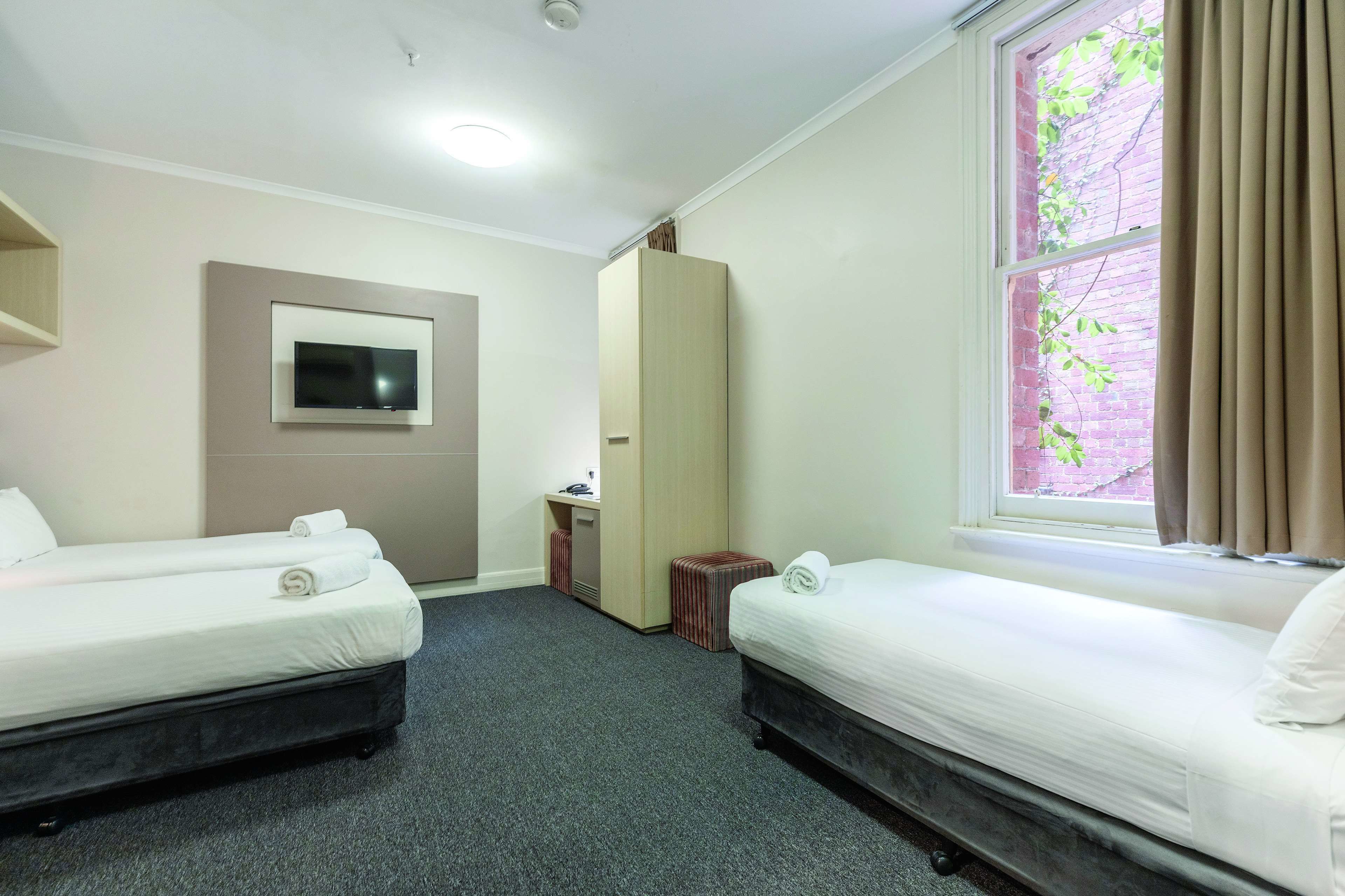 Habitación de hotel con dos camas, un televisor montado en la pared, un escritorio con una silla y una ventana con cortinas.