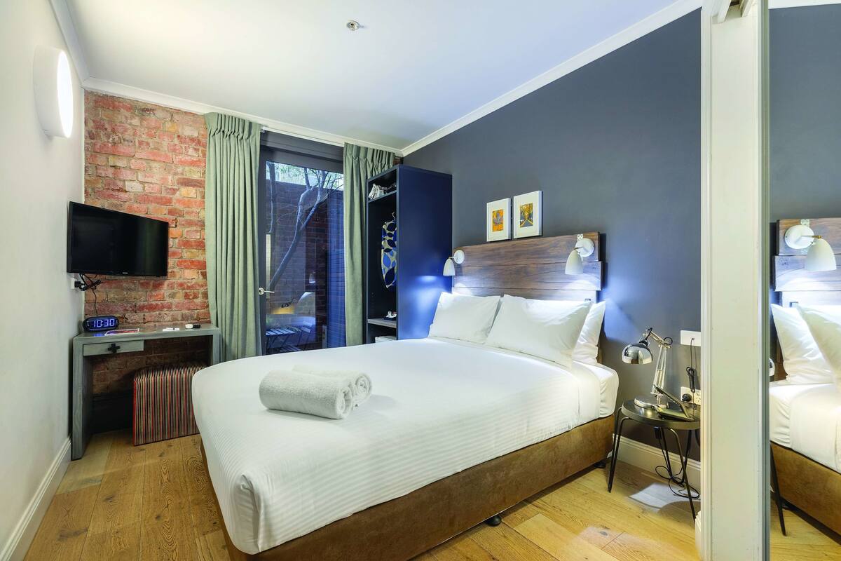 Una camera d'albergo moderna con un letto grande, una scrivania con televisore, una parete decorata con mattoni e vista attraverso una porta di vetro.