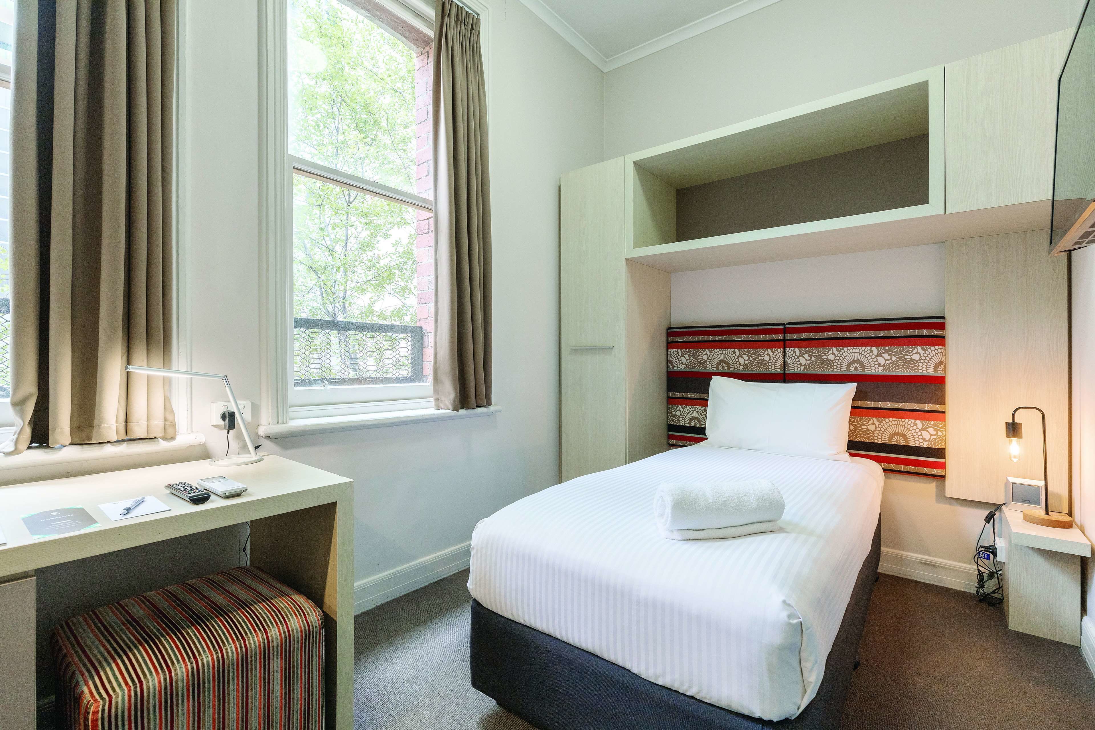 Ein Hotelzimmer mit Bett, Schreibtisch, Stuhl, einem Fenster mit Vorhängen sowie einem Kopfteil mit dekorativem Muster.