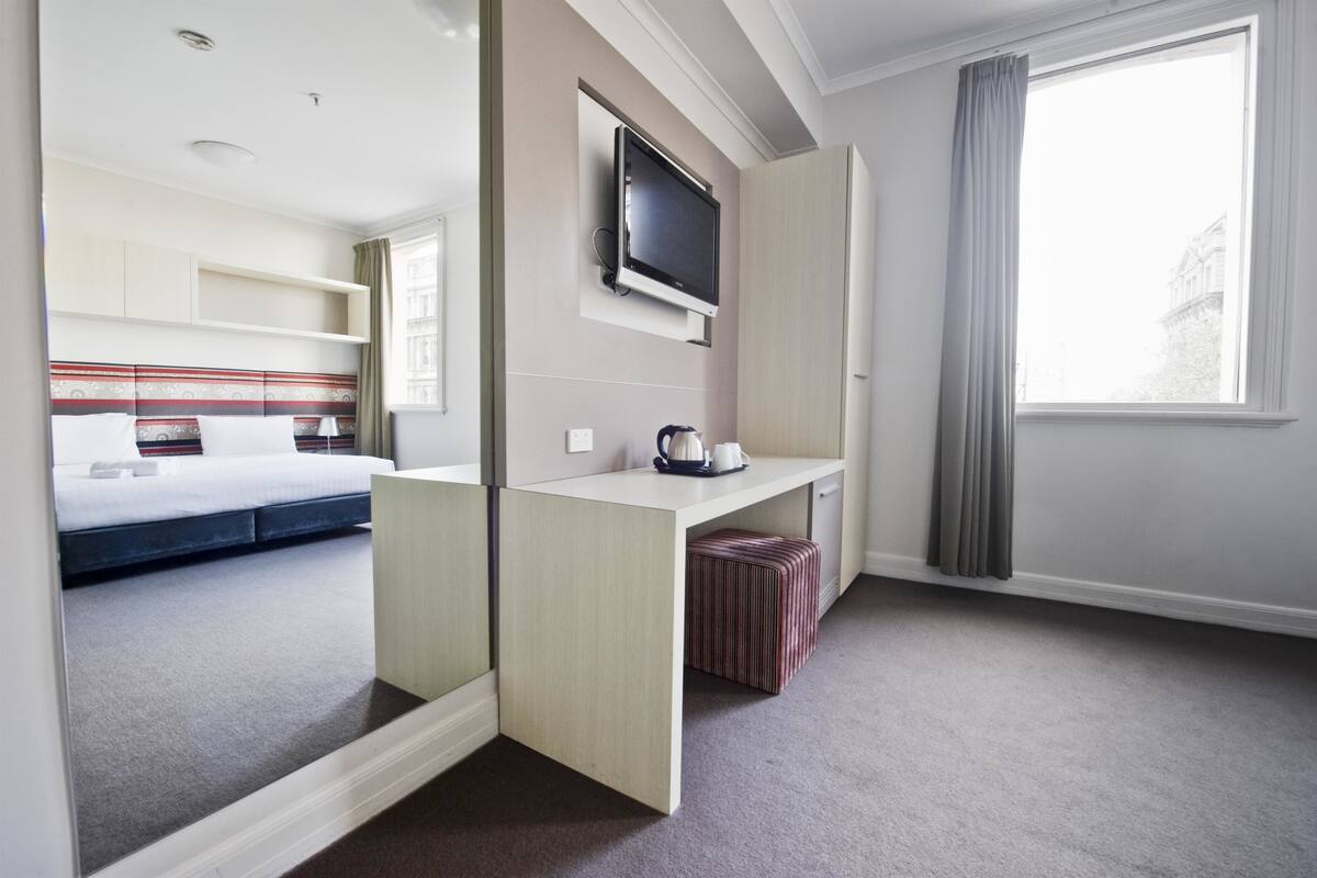 Una camera d'albergo con un letto, una scrivania con televisore, uno specchio e una finestra con tende.