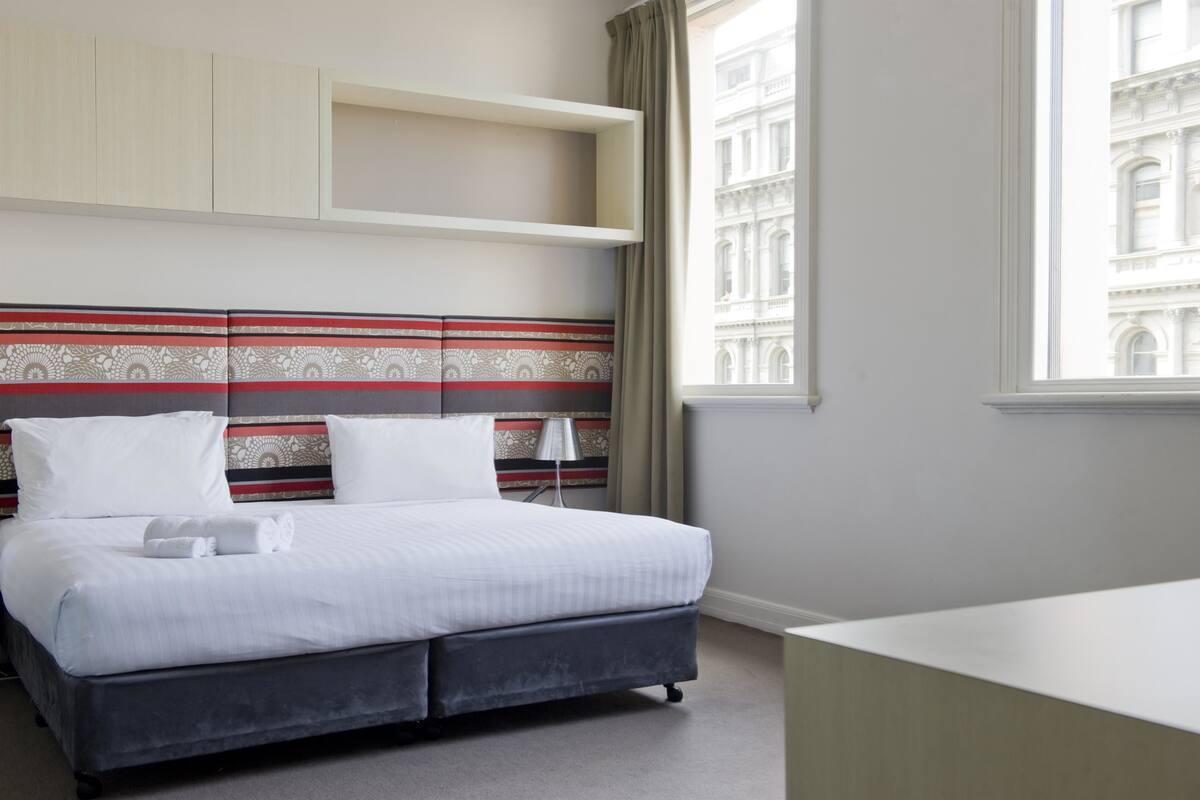 Una camera d'albergo con un letto, una scrivania, una lampada e una finestra che dà su un edificio.