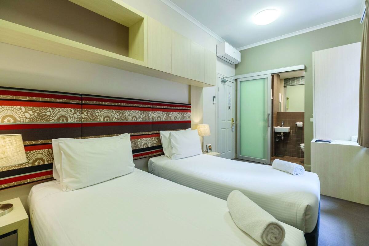 Una camera d'albergo con due letti, una testiera a motivi decorativi, un bagno con lavandino e specchio, e un condizionatore d'aria.