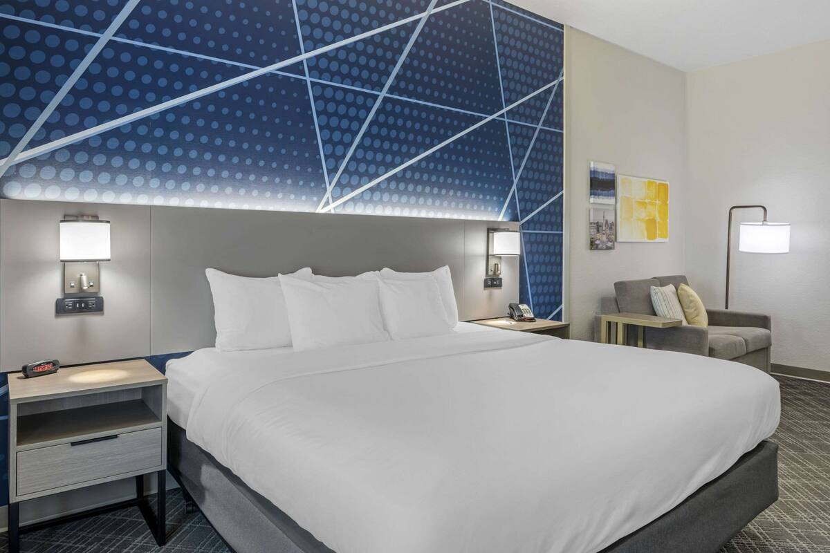 Habitación de hotel con una cama grande, una mesita de noche con lámpara, una silla y una pared con diseño geométrico.