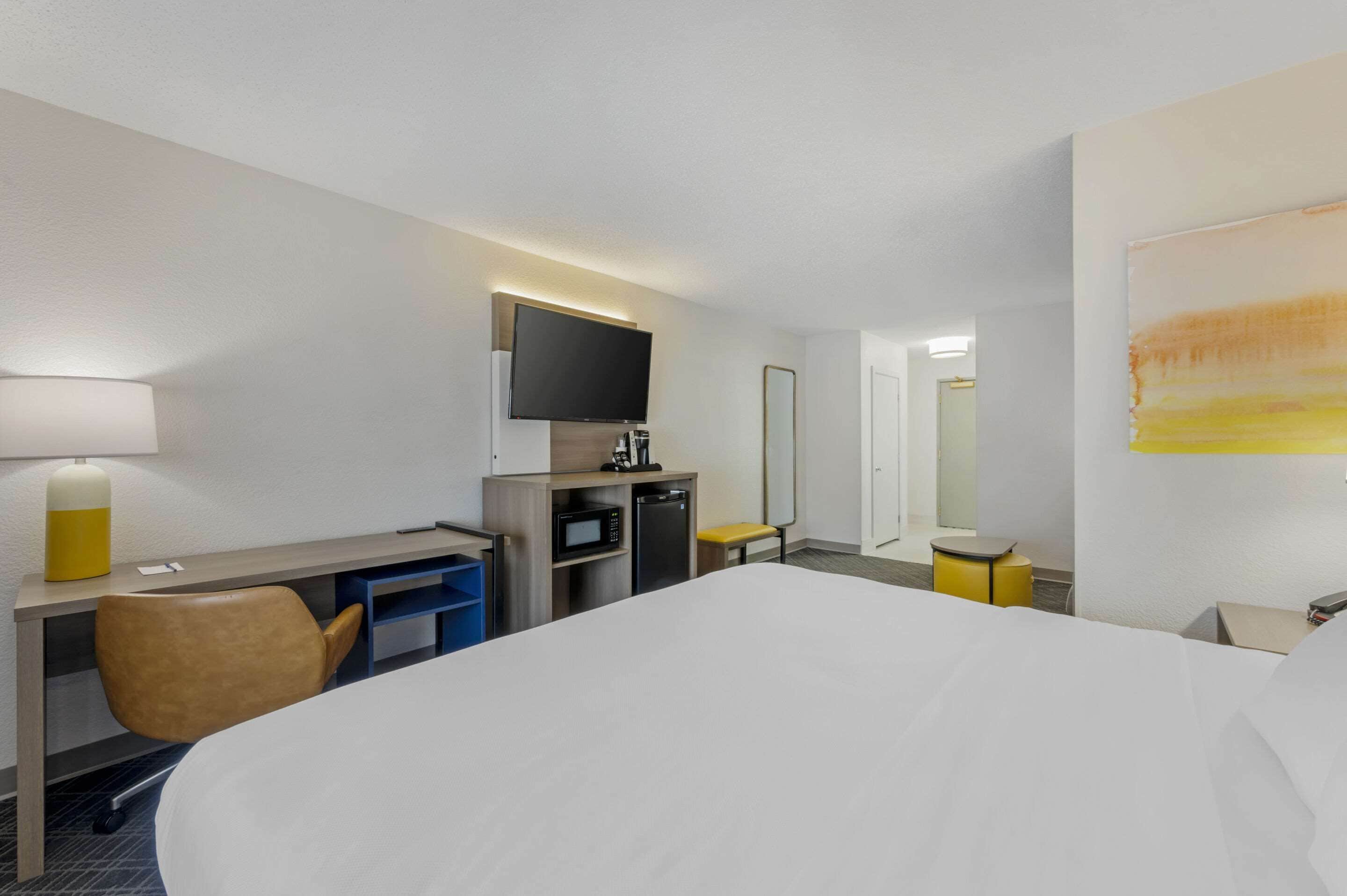 A hotel room with a large bed, a desk with a flat-screen TV, a microwave, a mini-fridge, a chair, and a small table.