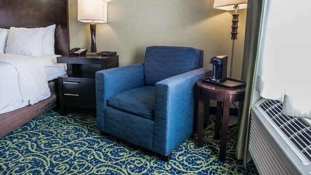 A hotel room with a bed, a blue armchair, a nightstand with a lamp, and a small table with a phone and a remote control.