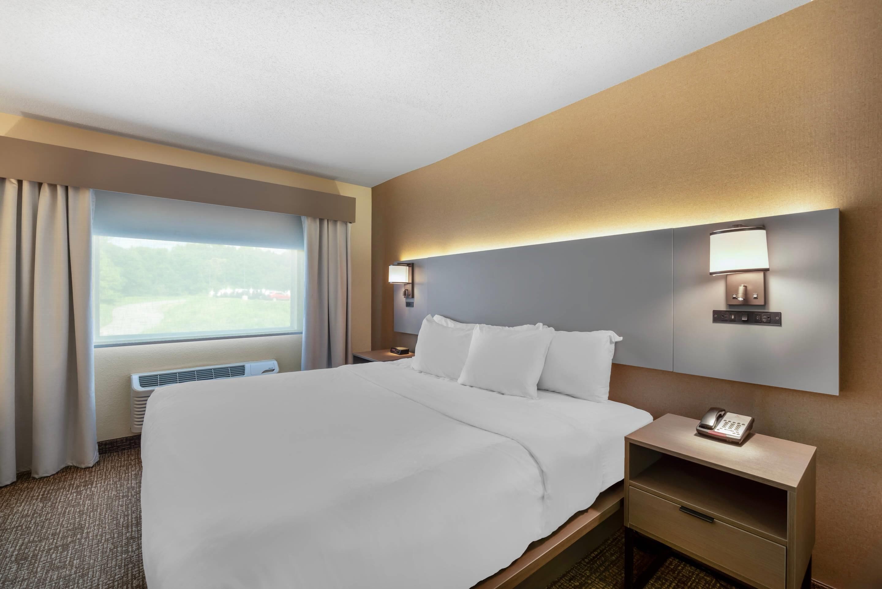 A hotel room with a large bed, a bedside table with a phone, a nightstand, a wall-mounted lamp, and a window with curtains.