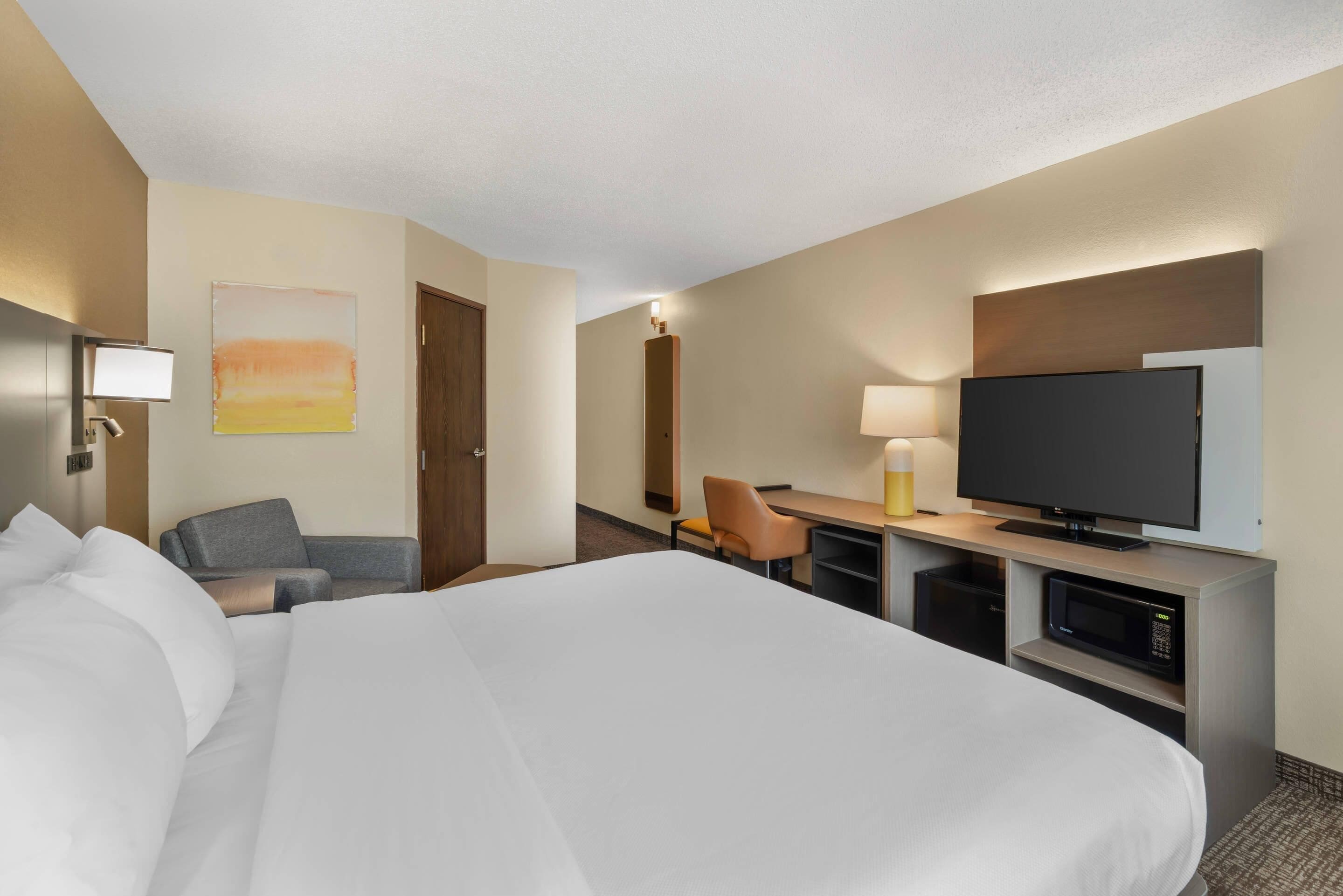 Habitación de hotel con una cama grande, un escritorio con televisor de pantalla plana, una silla y una mesita de noche con lámpara.