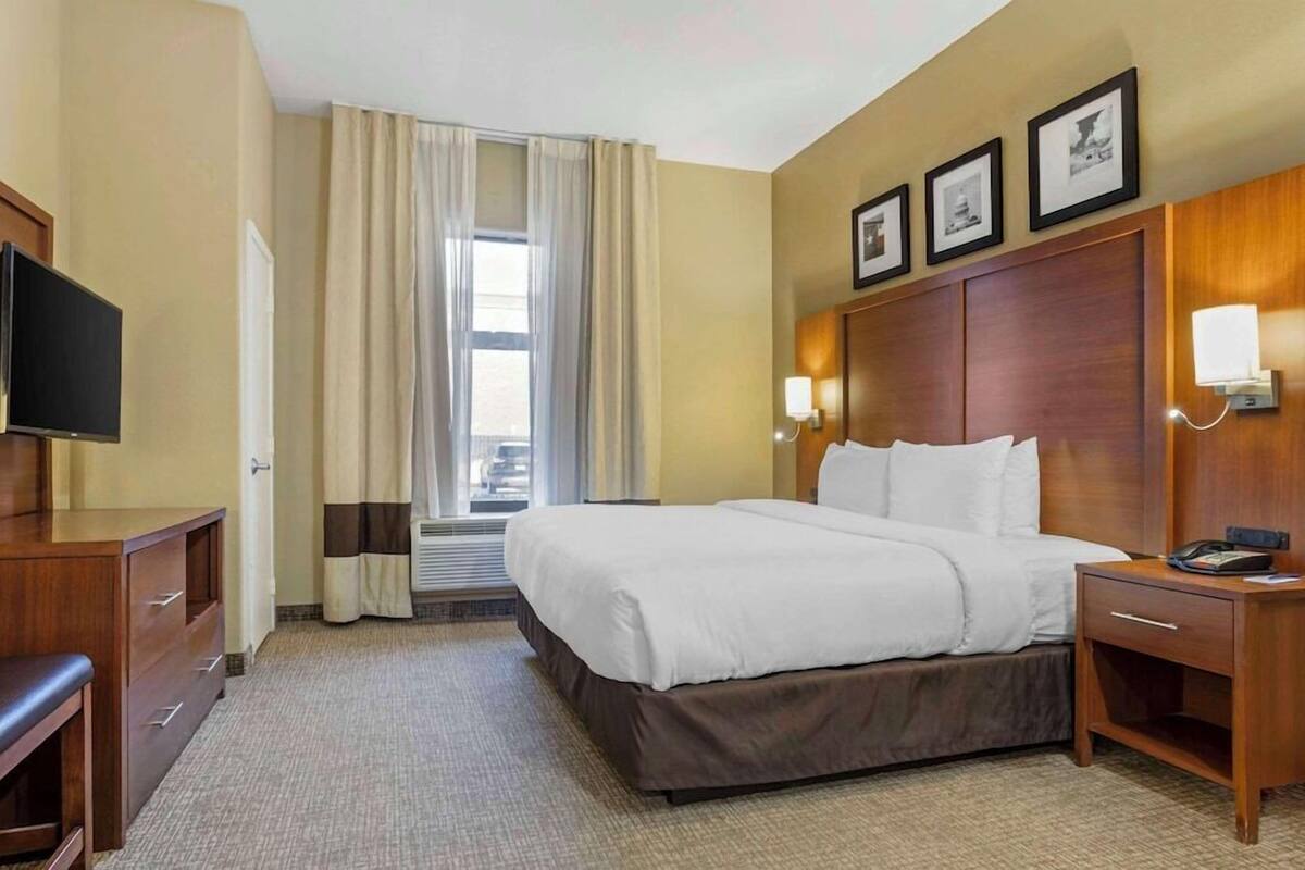 Une chambre d’hôtel avec un lit, un bureau, une télévision, une fenêtre avec des rideaux et des tableaux encadrés au mur.