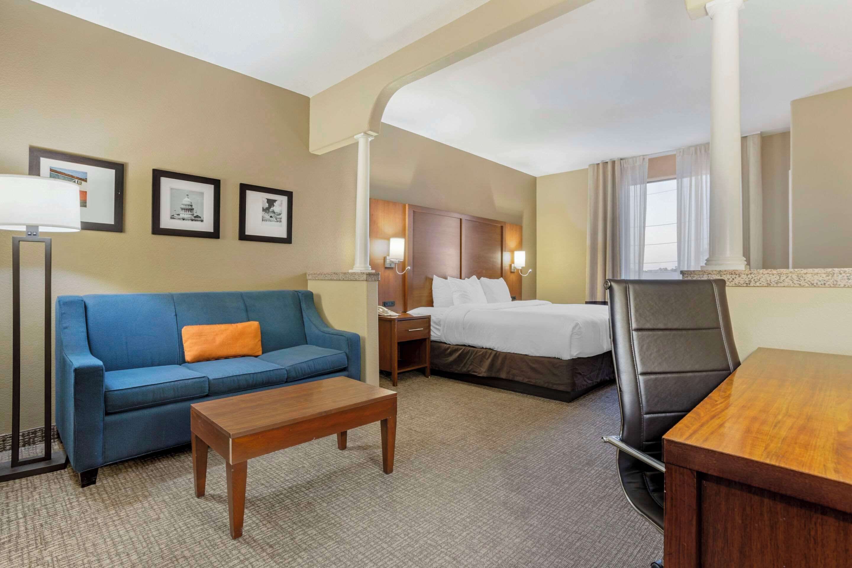 Une chambre d’hôtel avec un canapé bleu, une table en bois, un lit avec du linge de lit blanc, un bureau et une chaise. Les murs sont ornés de tableaux encadrés, il y a une table de chevet avec une lampe et une fenêtre avec des rideaux.