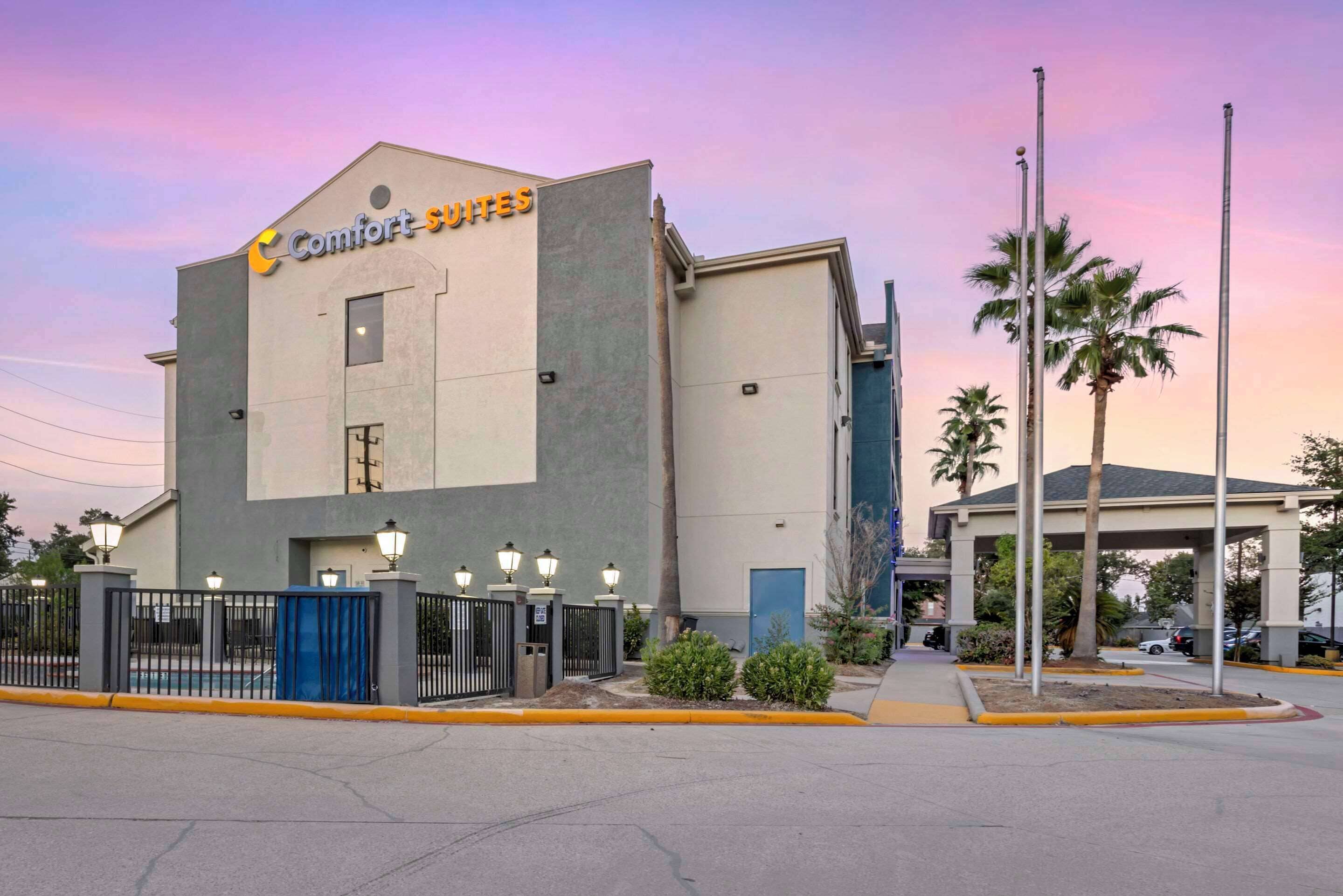 Un hotel Comfort Suites de diseño moderno, con entrada protegida por un portón, iluminación exterior y un estacionamiento con palmeras.
