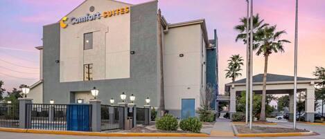 Un hotel Comfort Suites de diseño moderno, con entrada protegida por un portón, iluminación exterior y un estacionamiento con palmeras.
