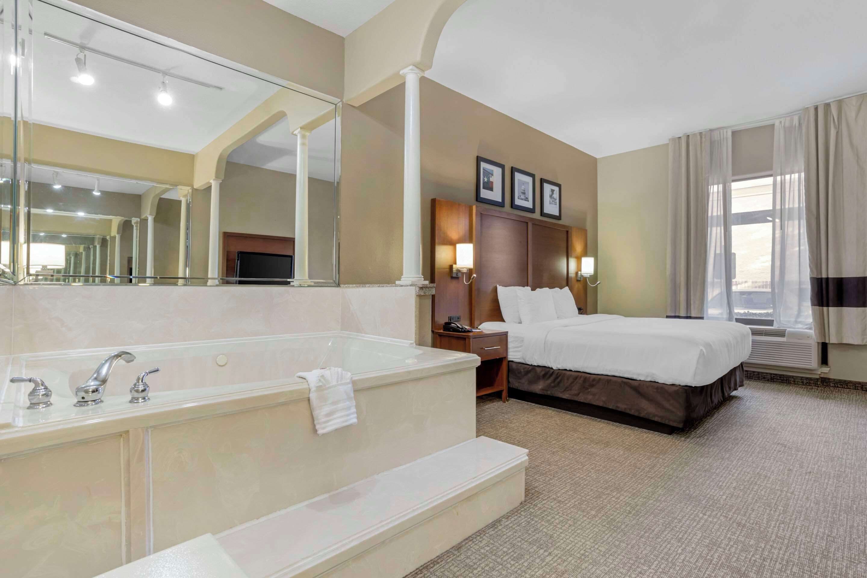 Een hotelkamer met een groot bad, een bed, een nachtkastje, een spiegel en een raam met gordijnen.