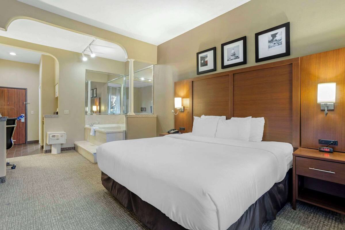 Une chambre d’hôtel avec un grand lit, une tête de lit en bois, une table de chevet, une commode, un miroir et une lampe fixée au mur.