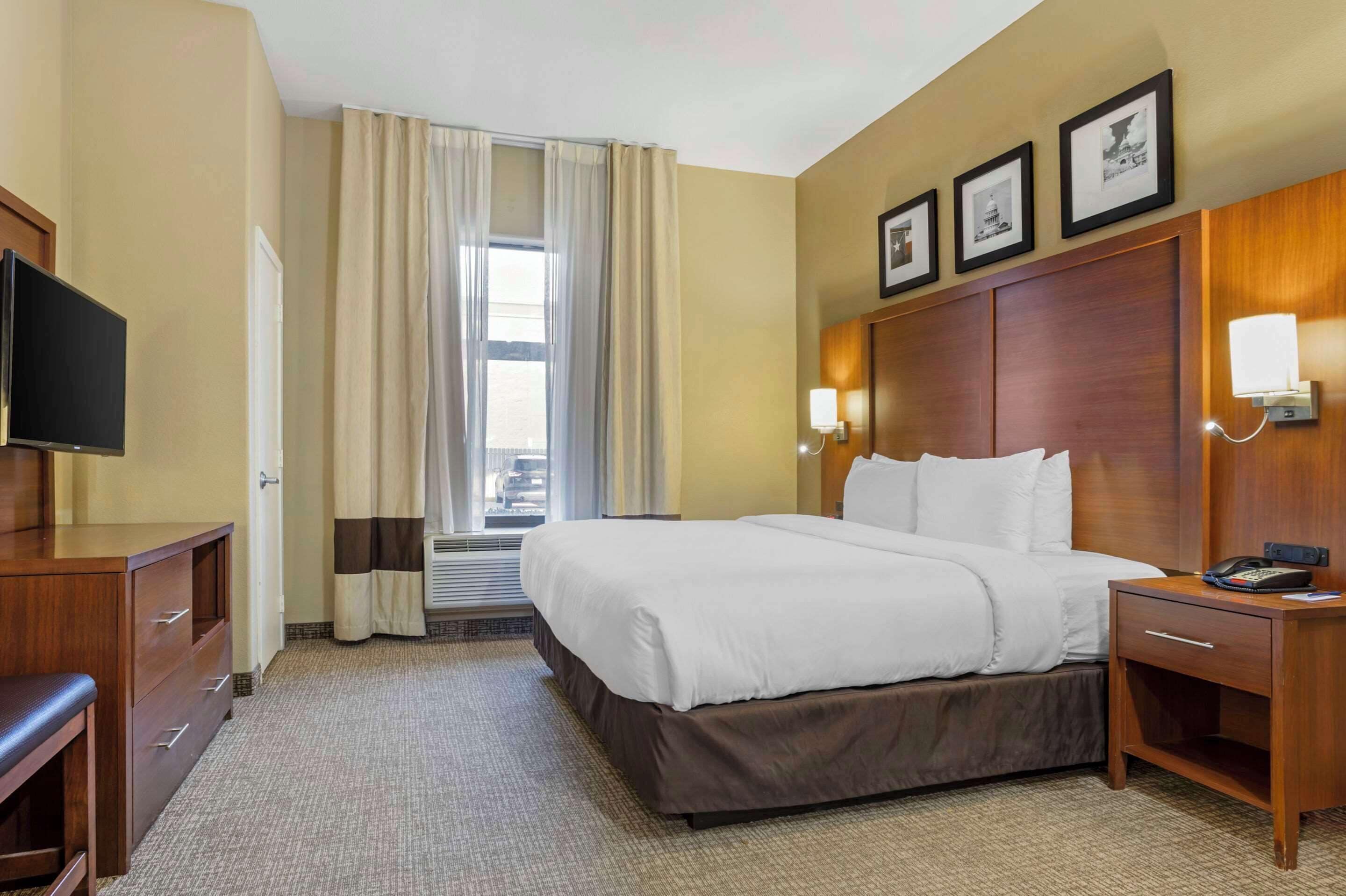 A hotel room with a bed, a desk, a television, a window with curtains, and framed pictures on the wall.