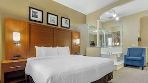 A hotel room with a large bed, a wooden headboard, a nightstand with a lamp, a blue armchair, a bathroom with a mirror, and framed pictures on the wall.