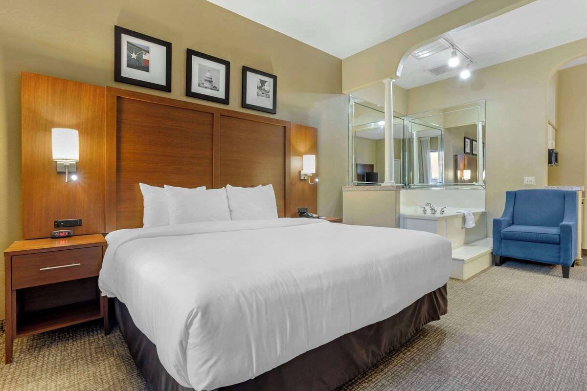 Une chambre d’hôtel avec un grand lit, une tête de lit en bois, une table de chevet avec une lampe, un fauteuil bleu, une salle de bain avec un miroir et des tableaux encadrés au mur.