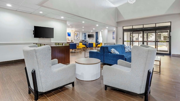 A spacious lobby with a high ceiling, a flat-screen TV mounted on the wall, a blue sofa, grey armchairs, a round coffee table, and a reception desk.