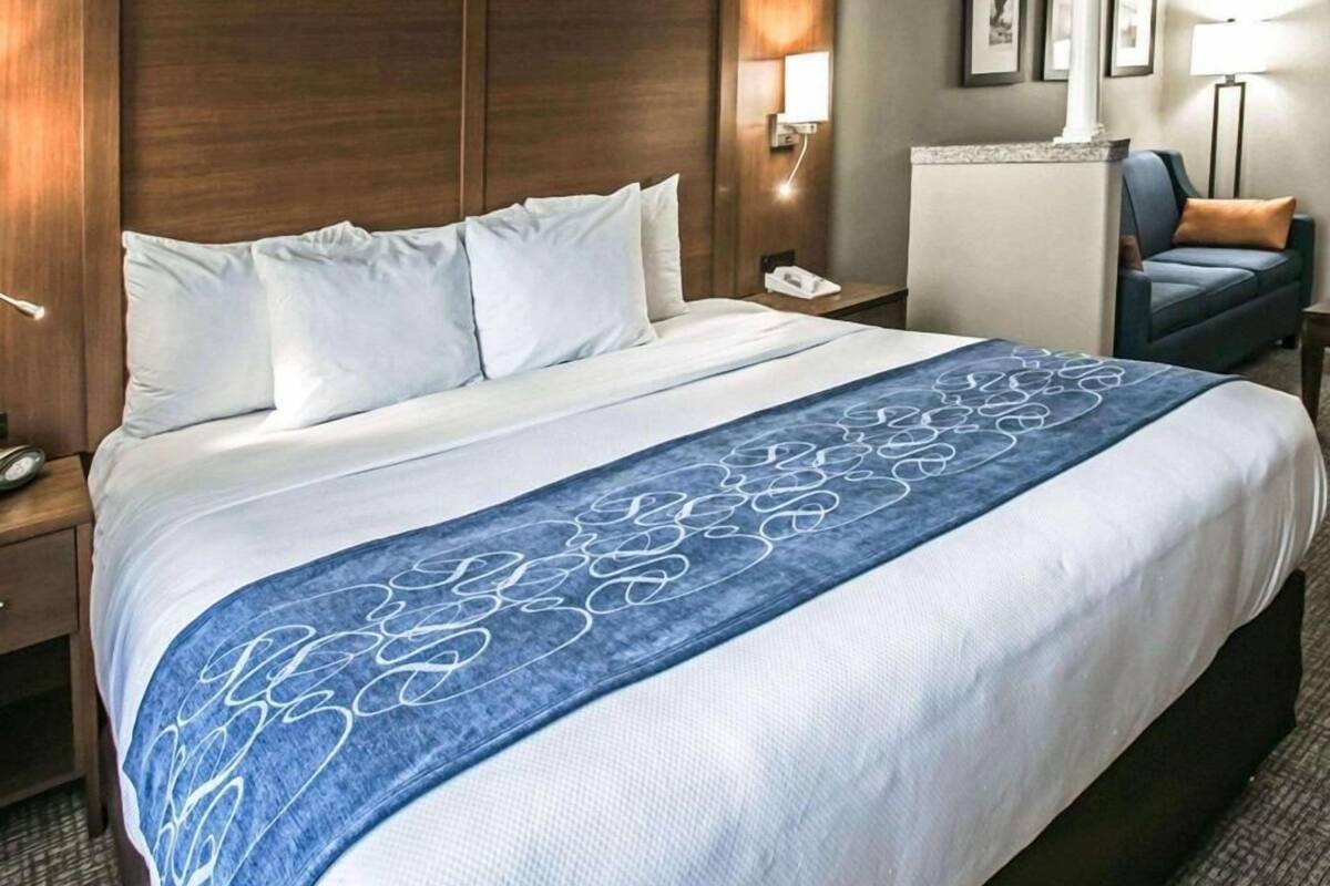 Une chambre d’hôtel avec un grand lit, une tête de lit en bois, une table de chevet avec un téléphone, une petite table de nuit, un bureau avec une lampe et une chaise avec un coussin.
