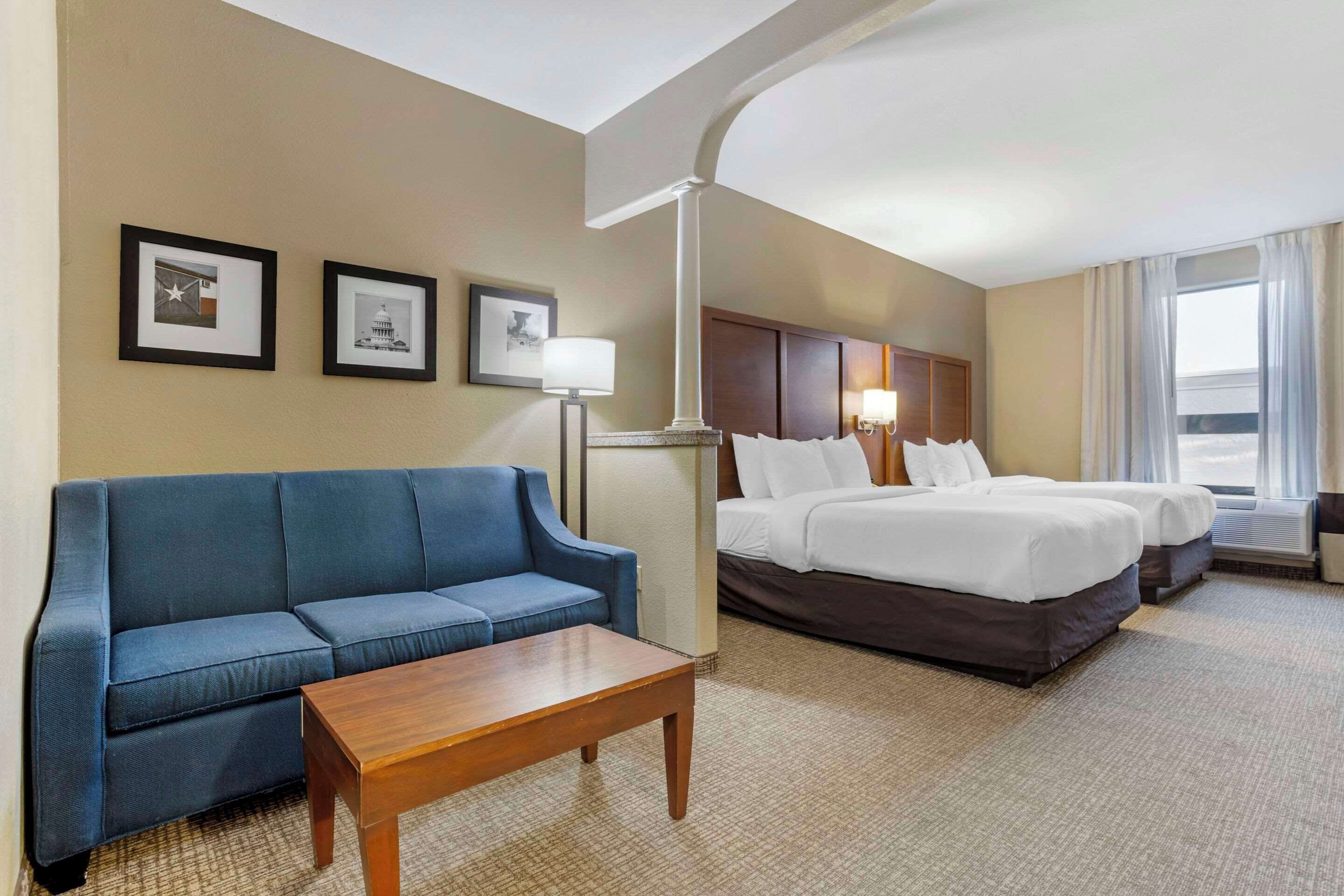 Habitación de hotel con un sofá azul, una mesa de centro de madera, dos camas con ropa de cama blanca y cuadros en la pared.