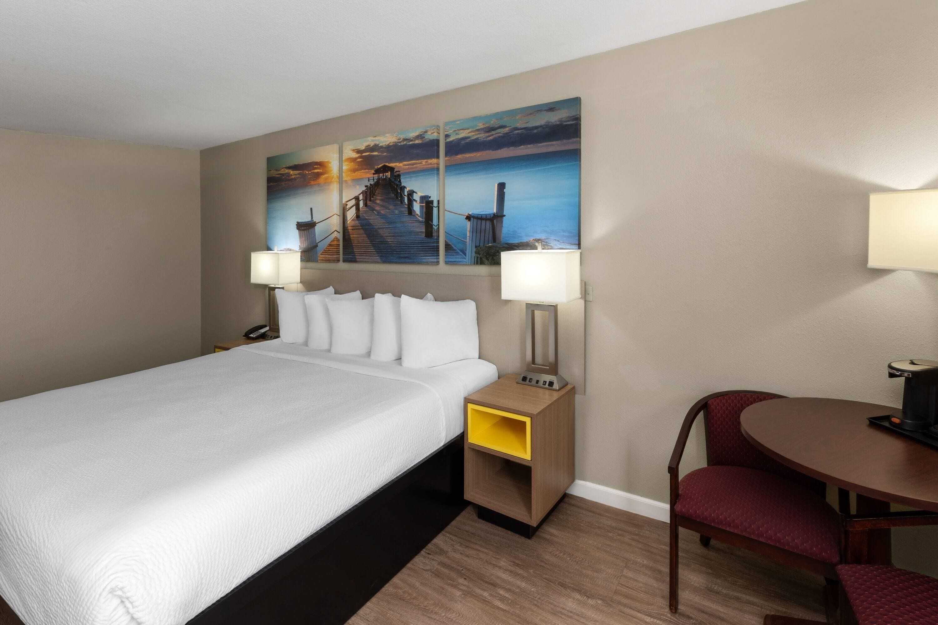 Ein Hotelzimmer mit einem großen Bett, einem Schreibtisch mit Lampe, einem Sessel und einem Gemälde eines Piers an der Wand.