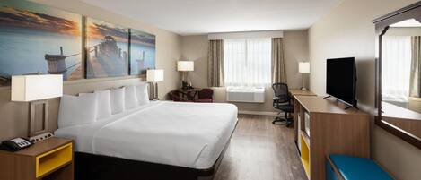A hotel room with a large bed, a desk with a TV, a chair, a desk lamp, a bedside table with a phone, and a painting of a pier on the wall.